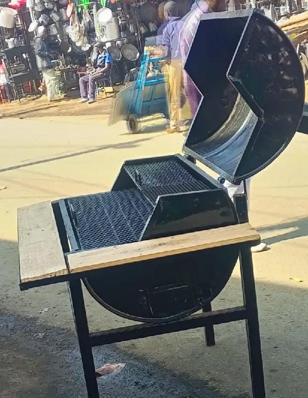 heavy-duty, charcoal-powered barbecue grill, often referred to as a "Juakali grill" in the local market. It is made from a repurposed metal drum and features a sturdy stand with wooden side tables. 
S