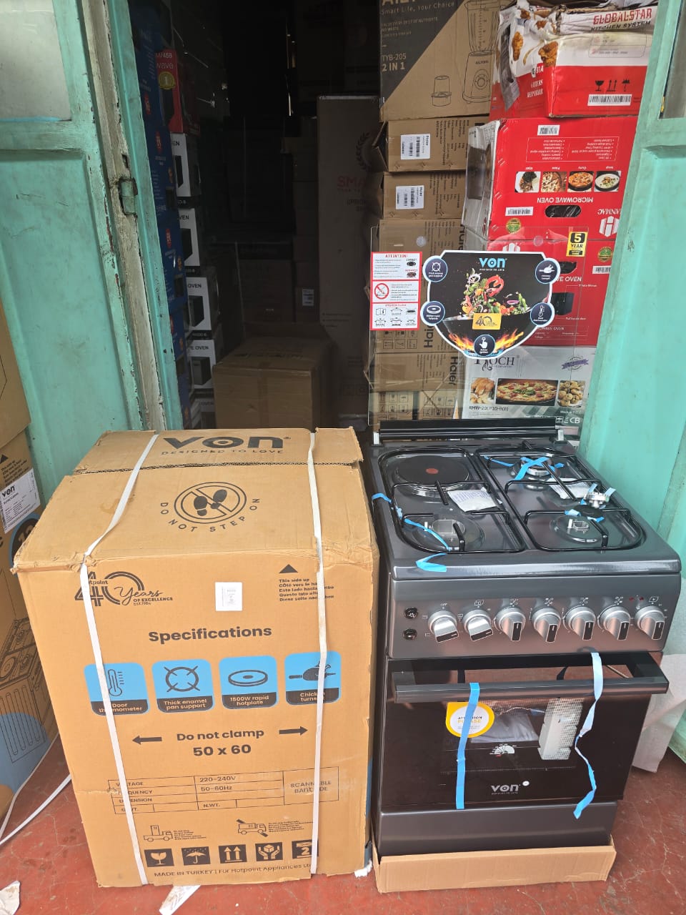 Von standing cooker and a boxed Von microwave oven in a retail setting.

The standing cooker is a gas model with four burners and an oven, likely featuring a matte finish and auto-ignition. 

The boxe