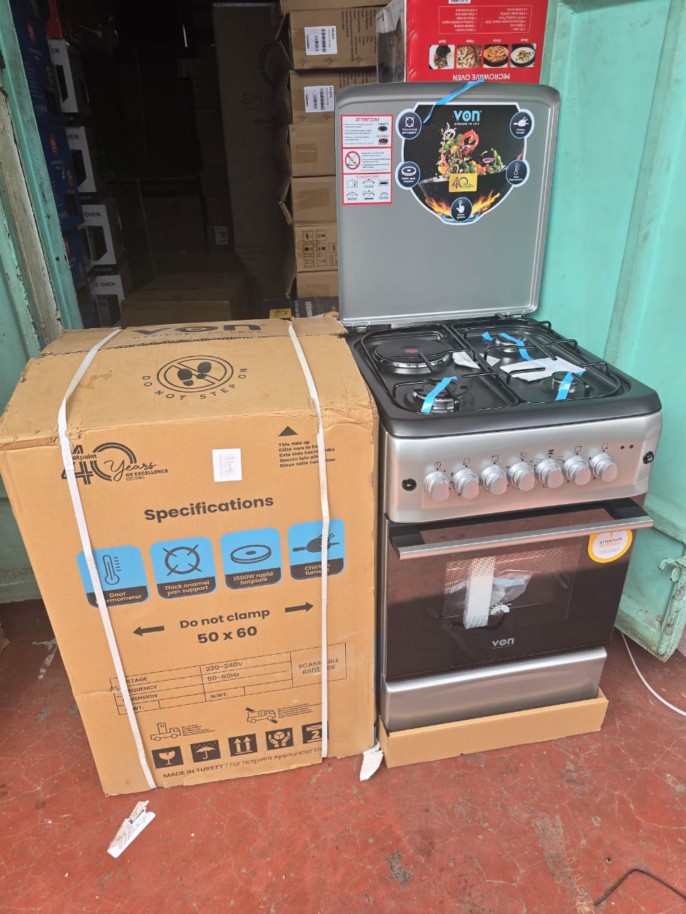 Von standing cooker and a boxed Von microwave oven in a retail setting.

The standing cooker is a gas model with four burners and an oven, likely featuring a matte finish and auto-ignition. 

The boxe